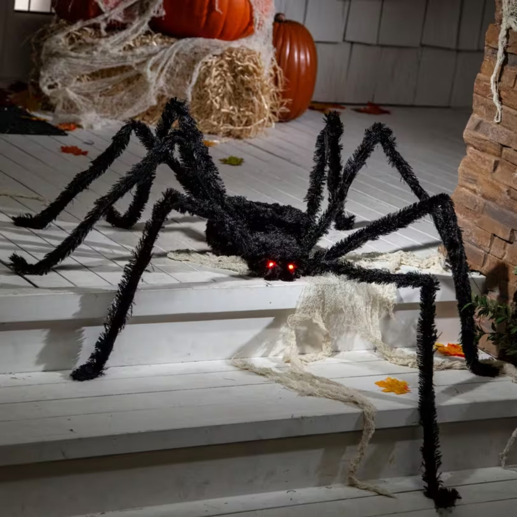 A large black fuzzy spider Halloween decoration with glowing red eyes sits on white porch steps, surrounded by cobwebs, straw bales, pumpkins, and autumn leaves.